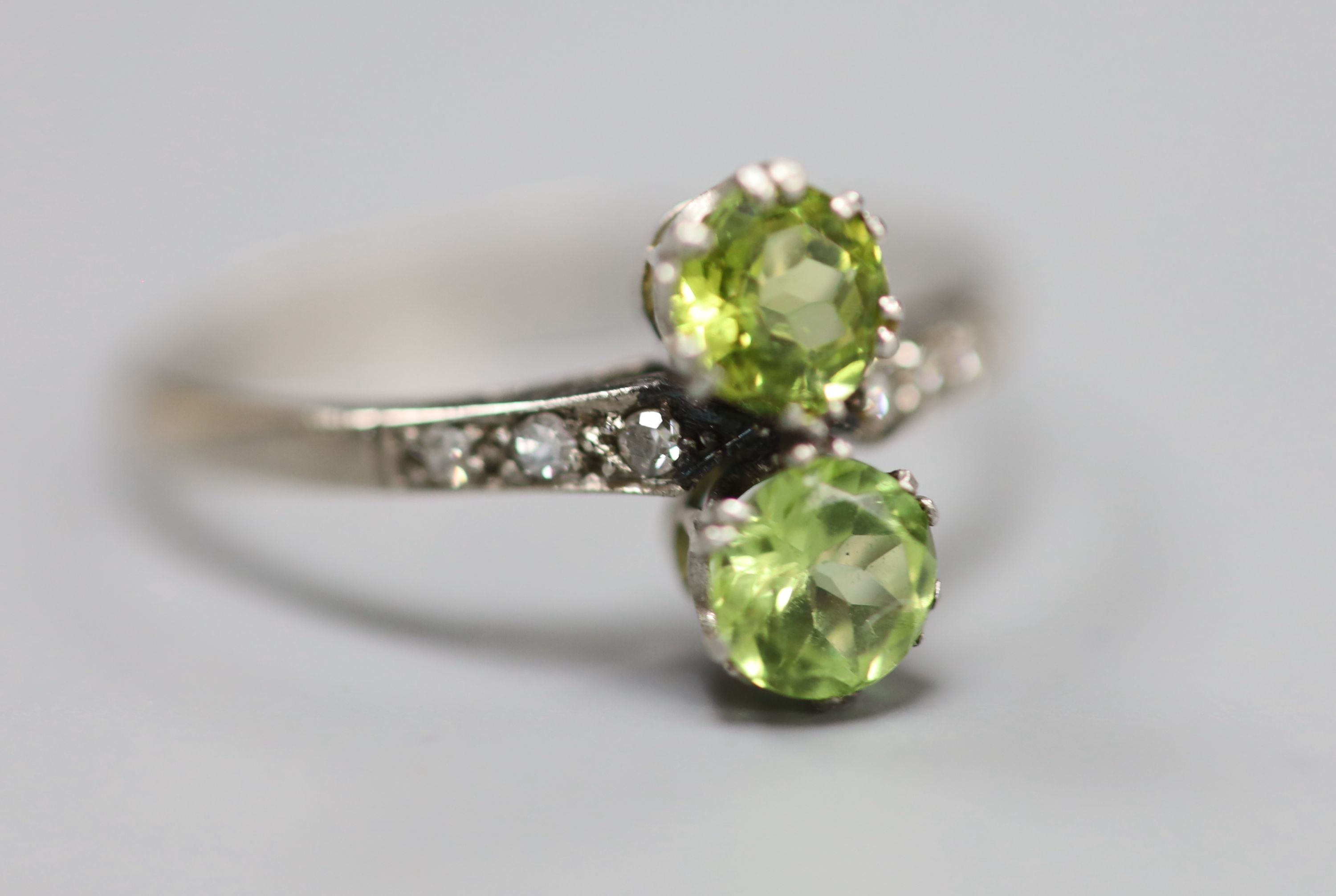 An 18ct and plat, two stone peridot crossover ring, with diamond set shoulders, size W, gross 2.7 grams.